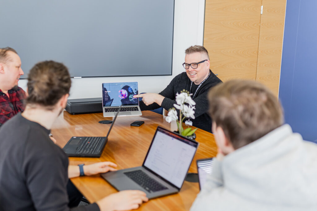 Mies esittämässä jotain tietokoneeltaan kolmelle miehelle pöydän äärellä