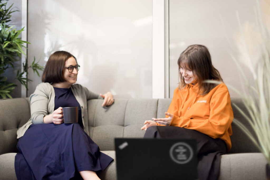Kaksi naista istuvat sohvalla keskustelemassa iloisesti kahvikupit käsissään